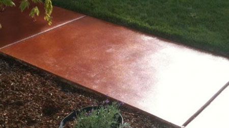 An outdoor concrete pathway wet from rain, divided into two sections with the nearer section having a reddish-brown tint and the farther section a natural gray. The path is bordered by lush green grass on one side and mulch with a potted plant on the other.