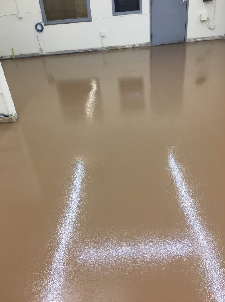 Interior view of an industrial space with a glossy tan floor showing reflections of white doors and windows, and bright white lines from overhead lighting.