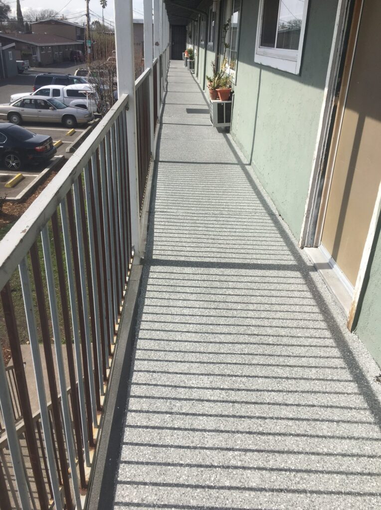Perspective view of an outdoor walkway along a building with a metal railing on one side, textured floor, and potted plants outside the doors; a parking lot with cars is visible on the left.