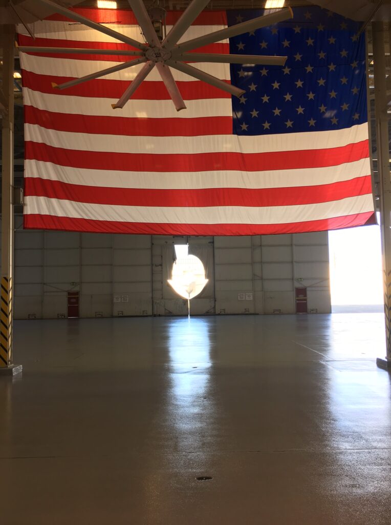 Large American flag hanging from the ceiling in a spacious industrial hangar, with a big ceiling fan above and sunlight streaming in from a doorway, casting a bright reflection on the glossy concrete floor.