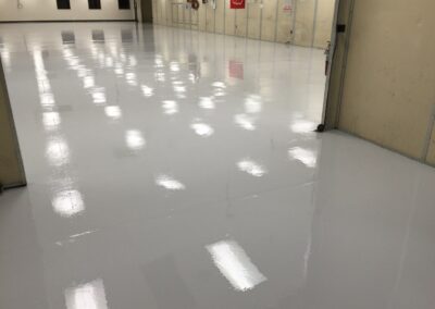 A large industrial or commercial space with a shiny gray floor reflecting the ceiling lights. The periphery of the room features beige walls with several doors and fire extinguisher stations, indicative of a focus on safety. The space appears clean, well-maintained, and possibly used for storage or as a work area.