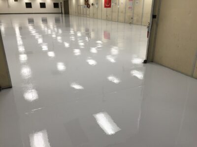 A large industrial or commercial space with a shiny gray floor reflecting the ceiling lights. The periphery of the room features beige walls with several doors and fire extinguisher stations, indicative of a focus on safety. The space appears clean, well-maintained, and possibly used for storage or as a work area.