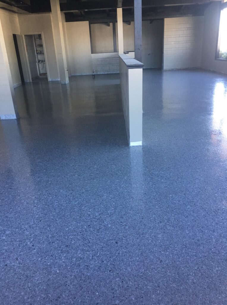 Open interior space with a speckled epoxy floor in shades of gray, complemented by block walls and structural columns. The area has a semi-industrial feel with visible utility lines running along the upper walls and natural light streaming in from windows on the far side.