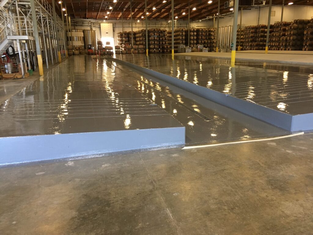 Large industrial warehouse with a glossy epoxy floor coating that reflects the bright ceiling lights. A prominent, long blue ramp runs through the space, providing access between different floor levels. Rows of stocked shelves with goods on pallets are visible in the background, highlighting the warehouse's storage function.