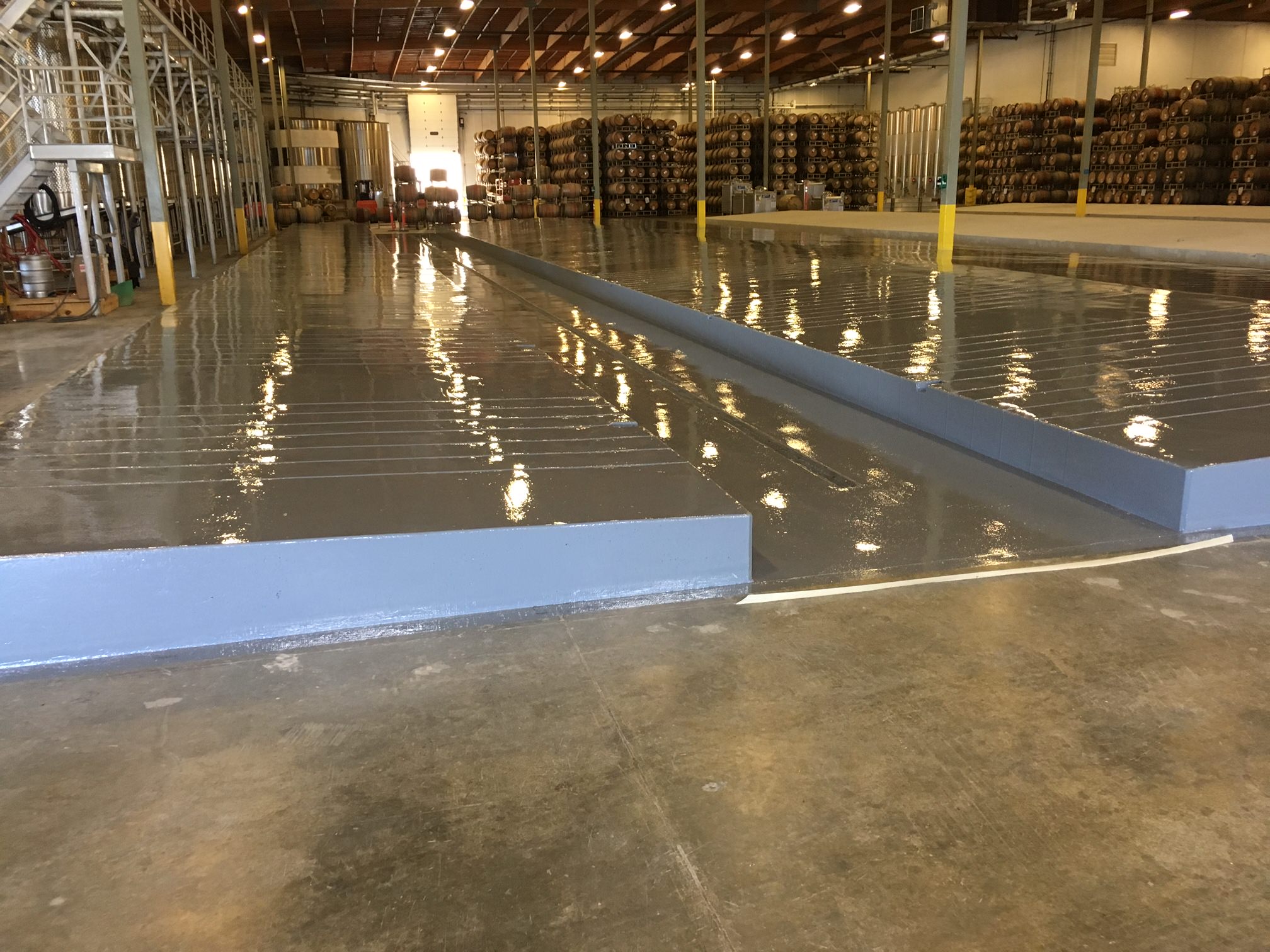 Large industrial warehouse with a glossy epoxy floor coating that reflects the bright ceiling lights. A prominent, long blue ramp runs through the space, providing access between different floor levels. Rows of stocked shelves with goods on pallets are visible in the background, highlighting the warehouse's storage function.