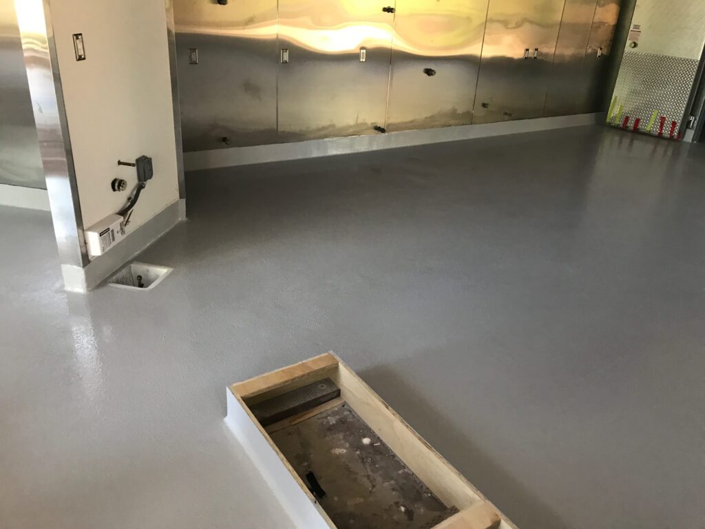 Industrial or commercial kitchen with a large, gray epoxy-coated floor. The kitchen features stainless steel wall panels and an exposed metal utility box. In the foreground, a wooden frame indicates a section under construction or repair, possibly a trench drain or access point.