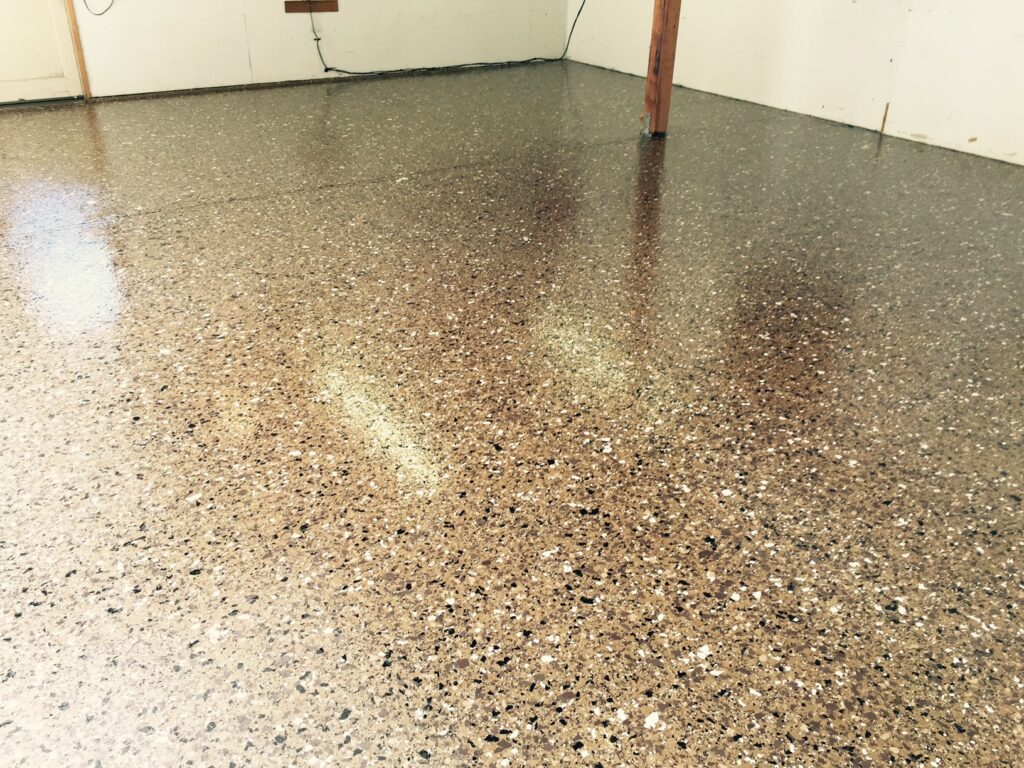 A room with a high-gloss epoxy floor, densely speckled in shades of brown, beige, and black. The light reflects off the smooth surface creating a warm glow. In the background, there is a simple wooden post and white walls with some scuff marks, indicating the space is used for practical purposes, possibly a workshop or a garage.