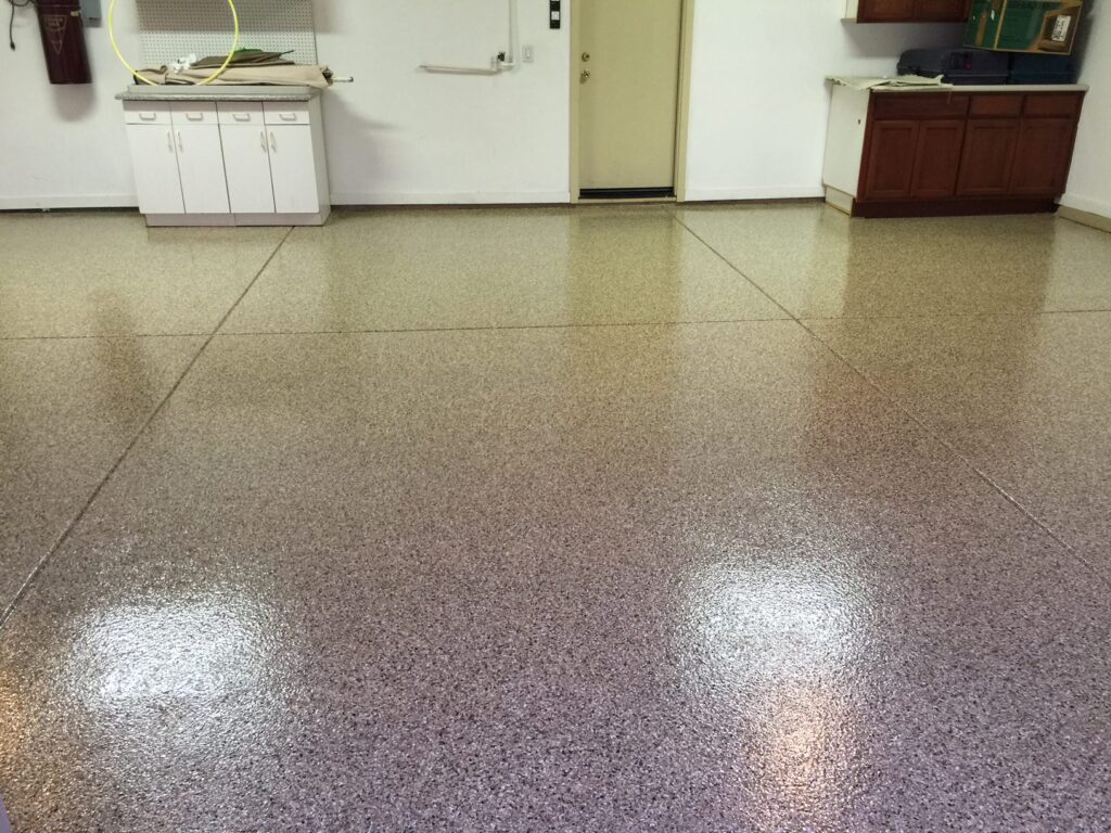 A room with tan and mauve speckled epoxy flooring, reflecting overhead lighting. The room has a white cabinet with a countertop on the left, a dark wooden cabinet on the right, and a closed door in the center of the back wall. The space is clean and seems to be a work area or utility room, with various items on the countertops suggesting active use.