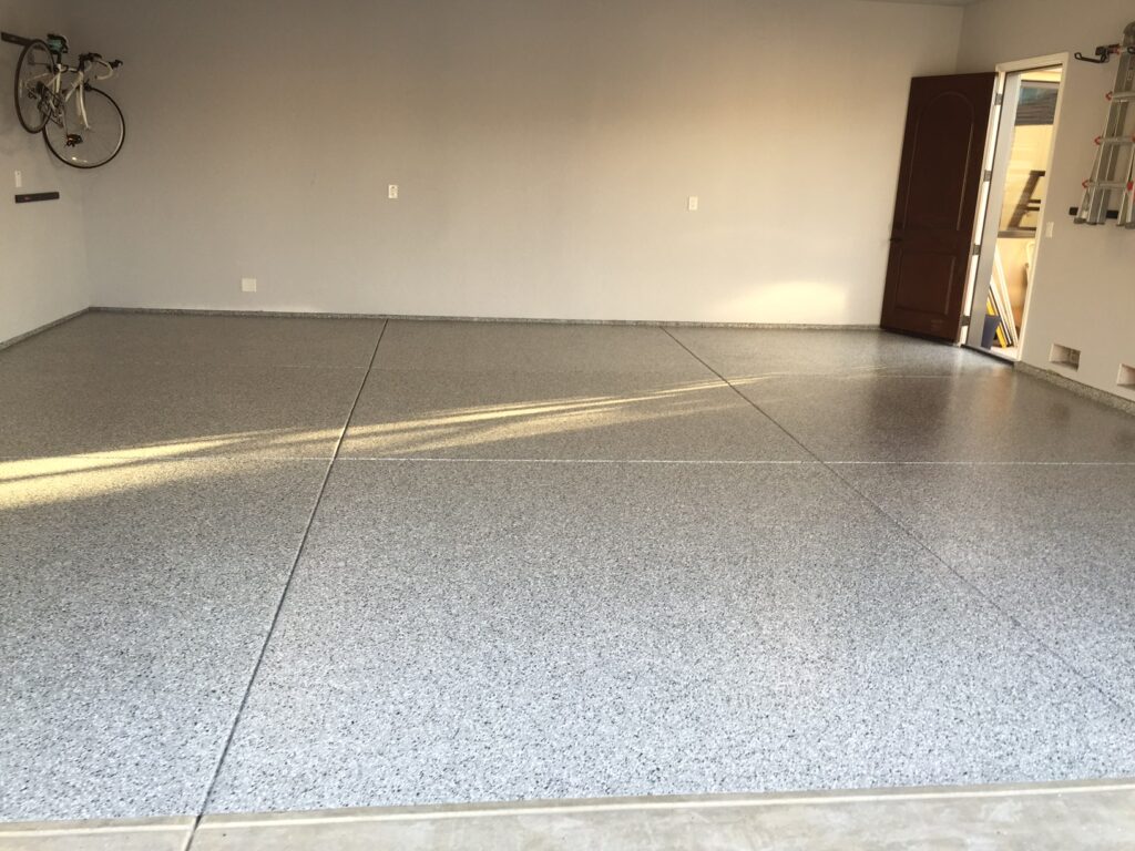 View from the driveway into a double garage with tan speckled epoxy flooring. The garage is open, revealing a clean, uncluttered space with two white doors on the back wall, a water heater to the right, and an electrical box on the left wall. The ceiling texture and walls are painted in a matching tan color, providing a uniform and tidy appearance.