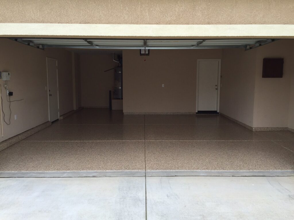 View from the driveway into a double garage with tan speckled epoxy flooring. The garage is open, revealing a clean, uncluttered space with two white doors on the back wall, a water heater to the right, and an electrical box on the left wall. The ceiling texture and walls are painted in a matching tan color, providing a uniform and tidy appearance.