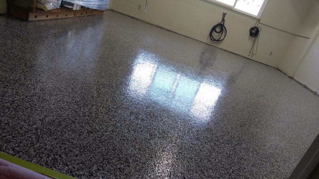 Interior of a room with glossy, speckled epoxy flooring reflecting natural light from windows. The space includes a beige wall with two coiled black cables hung neatly on the right. In the back left corner, a wooden pallet with items wrapped in clear plastic is partially visible. The area conveys a tidy, utilitarian atmosphere with ample lighting.