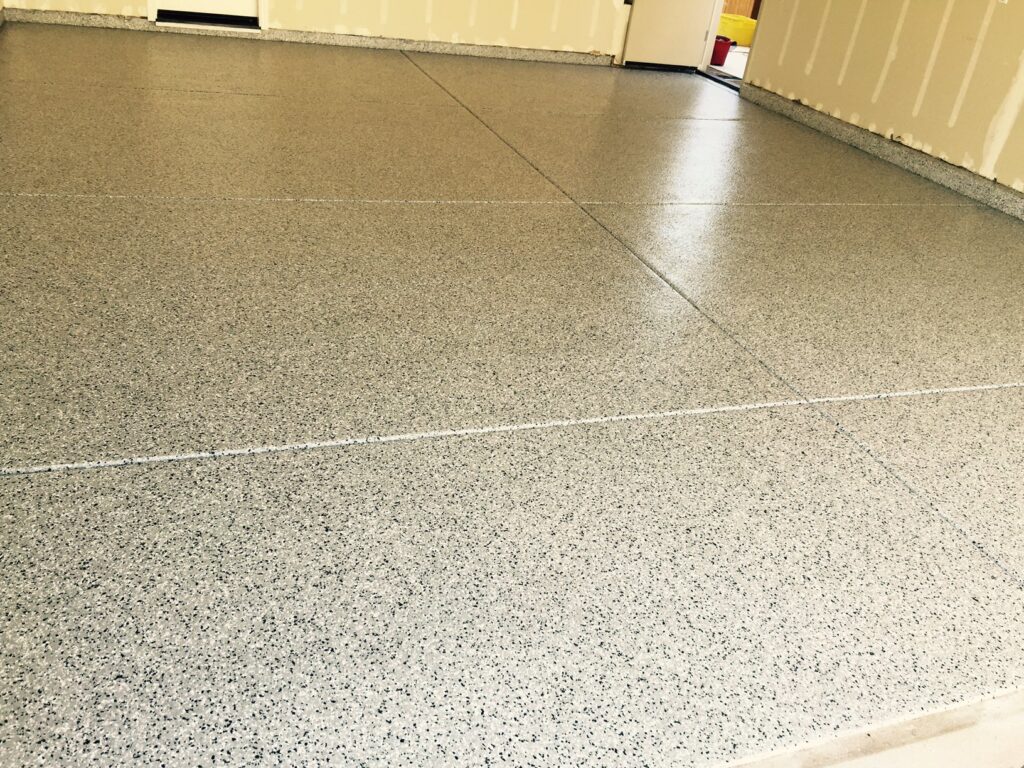 A close-up view of a room with a glossy speckled terrazzo floor. The cream-colored walls have a vent near the baseboard and an air conditioning unit on the lower wall to the left. The right corner of the room features a yellow safety pole next to a garage door, partially visible.