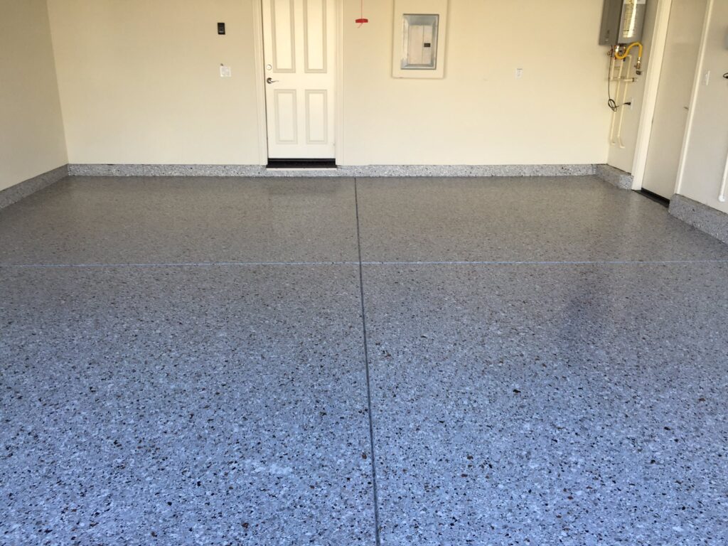 A clean, empty room with a light gray speckled epoxy floor. There are two white doors on the back wall, one closed and the other slightly open. To the right, there's a water heater and gas pipeline. The room also features a small white electrical panel on the right wall. The space looks like a functional area, possibly a laundry or utility room in a residential home.