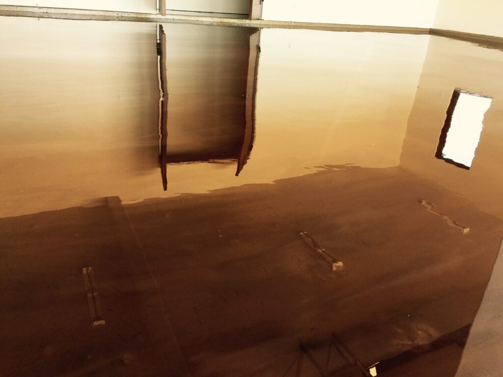 A large, empty room with a high-shine caramel-colored epoxy floor reflecting the ceiling's structure and a bright window. The reflections create a mirrored effect, giving the impression of a vast and open space. The smooth surface of the floor and its reflective quality indicate a modern and well-maintained interior, possibly in a commercial or industrial setting.