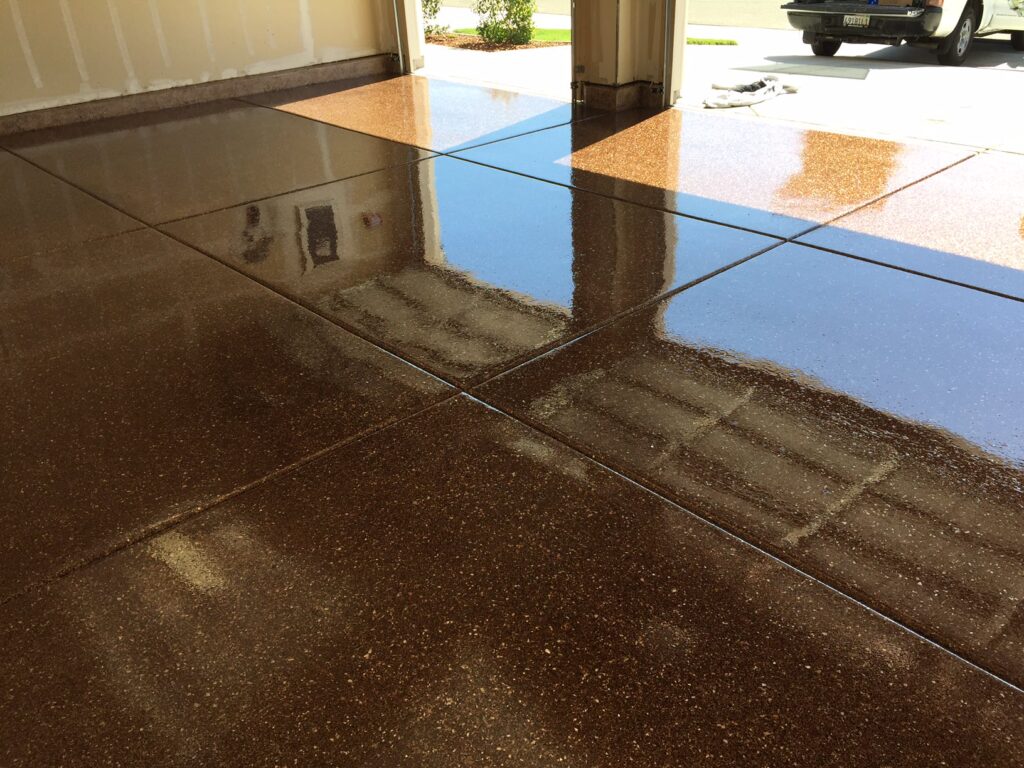 The image showcases a polished, sparkling brown decorative concrete epoxy floor of a garage with a high-gloss finish that reflects the surrounding walls and objects, including a vehicle parked outside. The open garage door lets in natural light, which enhances the reflective quality of the floor, showcasing its pristine condition and the detailed texture of the speckled surface.