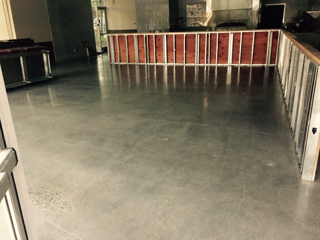 A view of an indoor space with a matte gray concrete floor, featuring a wooden and metal railing partitioning the area. In the background, countertops and various objects suggest a commercial setting, possibly a restaurant or bar under construction or renovation. The subdued lighting and visible construction materials convey an atmosphere of development and transition.