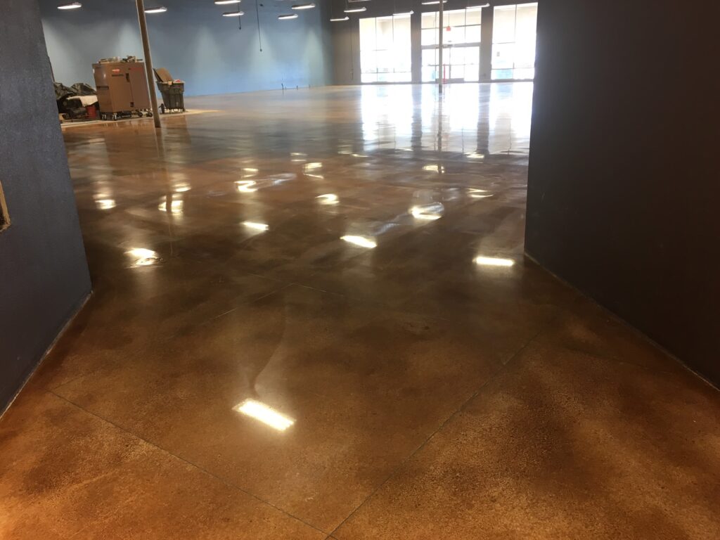 Expansive room with a polished concrete floor in a warm, amber hue, reflecting overhead lights, with large windows in the background and some equipment stored at the periphery, suggesting a commercial or industrial setting.