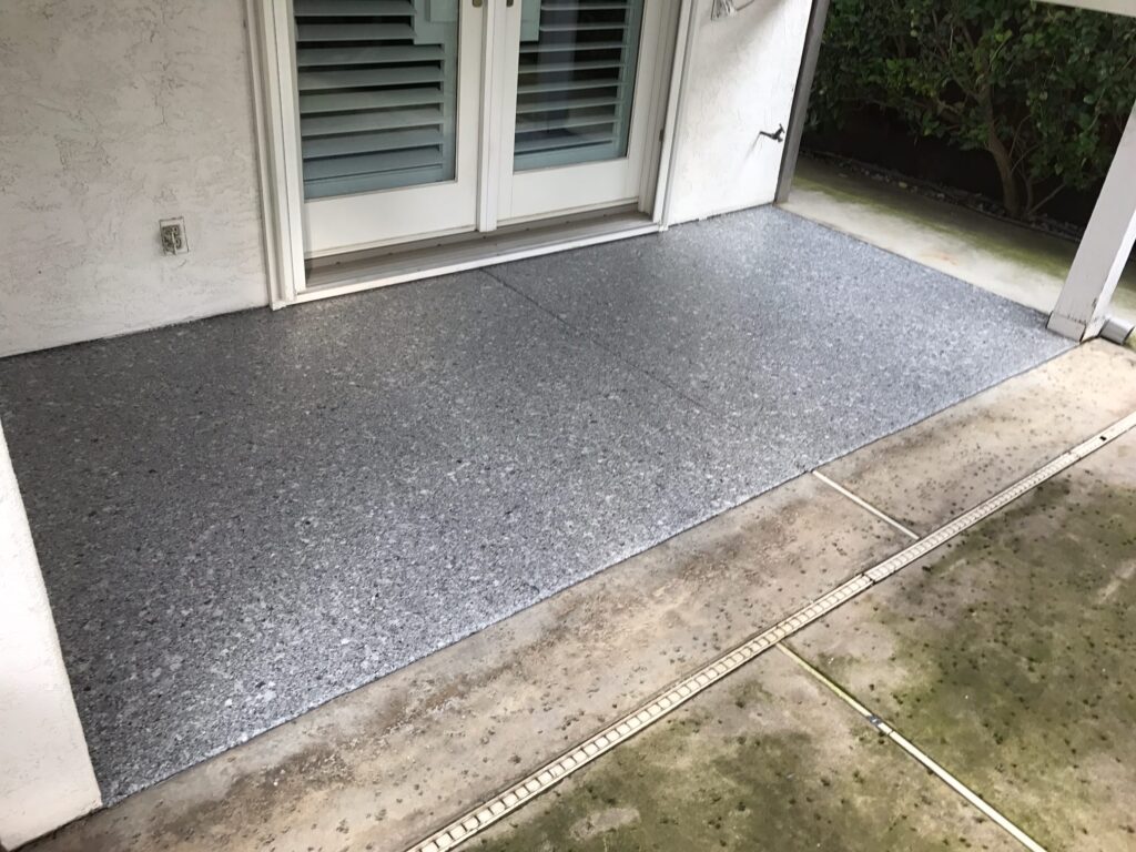 A residential outdoor entryway featuring a textured gray epoxy floor. It contrasts with the white stucco wall and a set of double doors with white frames and green shutters. The surrounding area includes a concrete path and a small garden bed, giving a welcoming approach to the home.