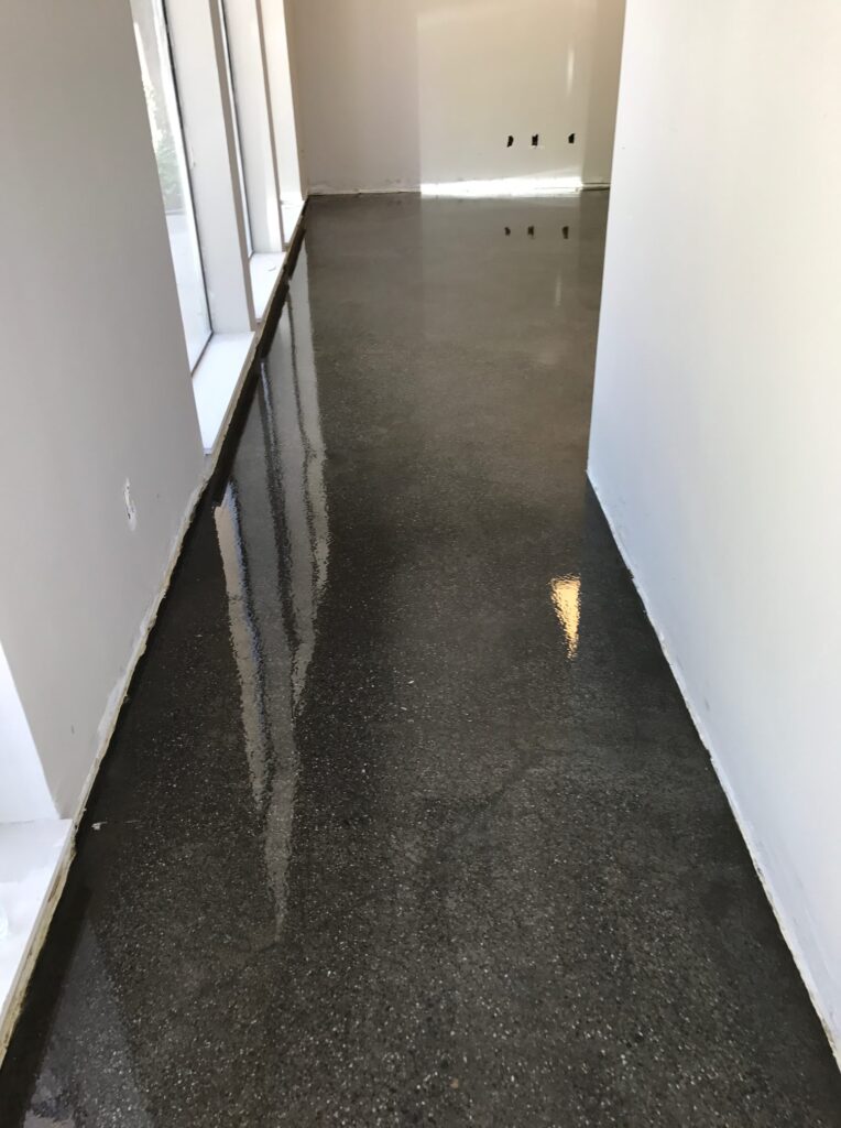 A narrow corridor with a high-gloss finished floor reflecting the natural light from floor-to-ceiling windows on one side, with a bright, airy feel and outlets visible on the opposite wall, suggesting a modern interior setting.