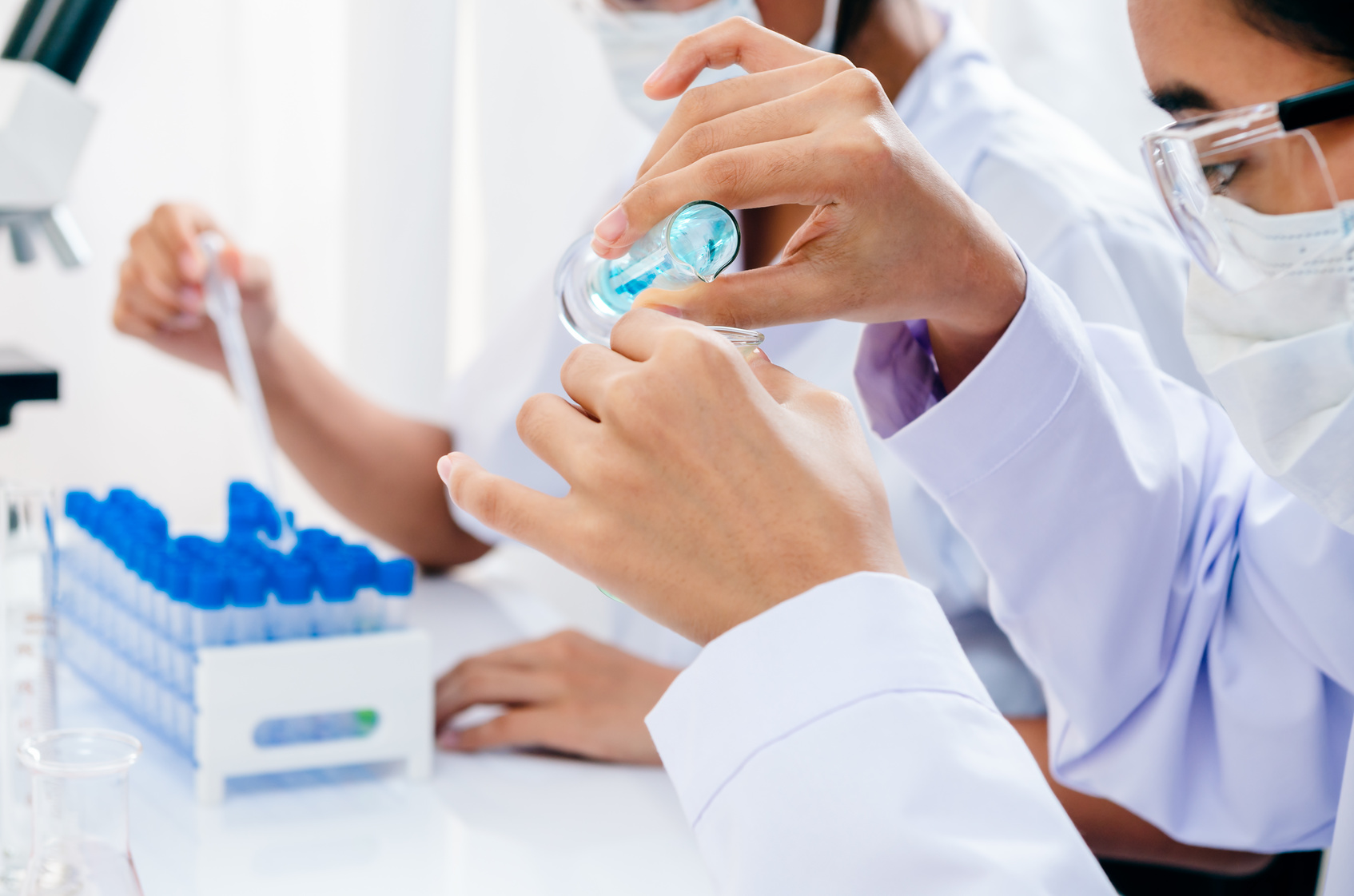 Close-up of two scientists in a laboratory, one handling a pipette and a test tube with a blue liquid. Both are wearing white lab coats, protective gloves, and safety glasses, adhering to safety protocols. The focus on their hands and equipment underscores the precision and concentration required in scientific research.