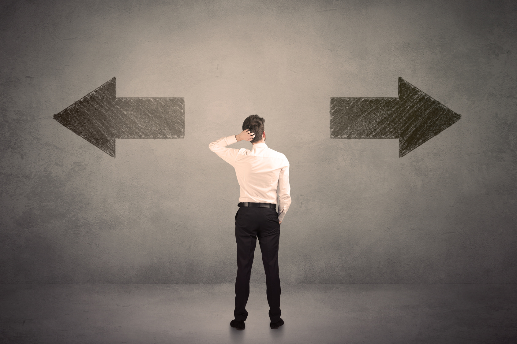 A man in business attire stands in front of a concrete wall, looking upwards and scratching his head in a gesture of contemplation or decision-making. Behind him, two large, chalk-drawn arrows point in opposite directions, symbolizing a crossroad or a need to make a choice between two different paths or options.