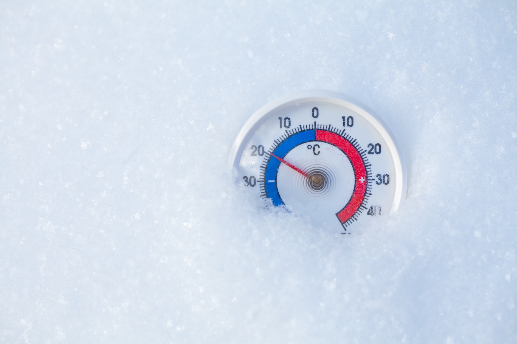 A round outdoor thermometer partially embedded in fresh snow, with the needle indicating a temperature around the freezing point. The thermometer has a clear, legible scale with red and blue color coding for temperature degrees in Celsius, providing a visual contrast against the white snowy background.