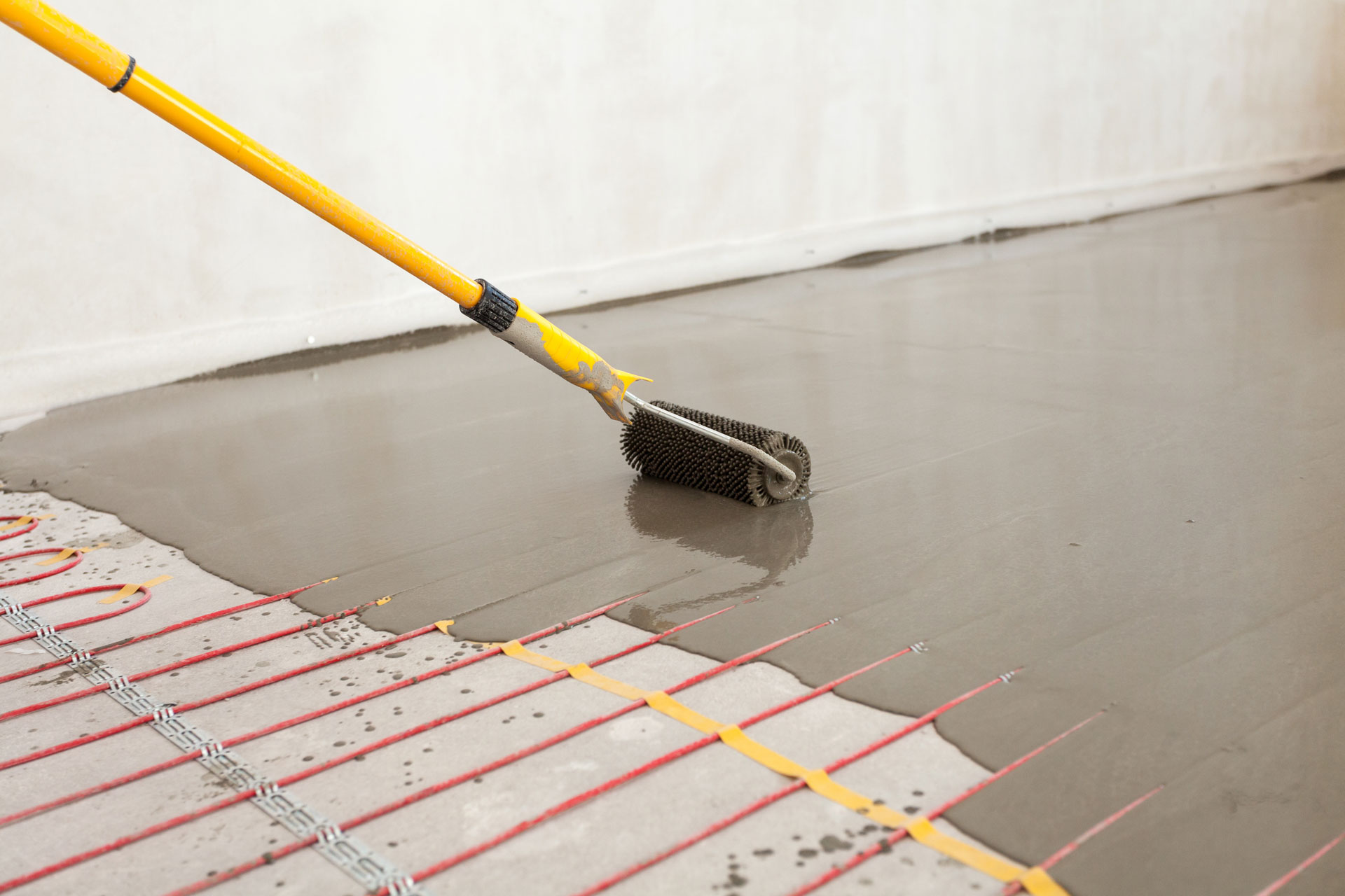 Application of a self-leveling compound on a concrete floor using a spiked roller attached to a yellow pole. The floor has underfloor heating pipes laid out in a grid pattern, visible through the semi-liquid layer of the compound. This image represents a stage in preparing a floor for final surface installation.