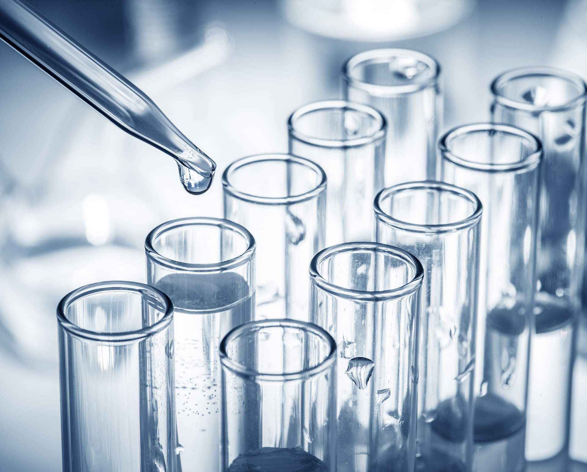 Close-up of a pipette dispensing a liquid droplet into a test tube among a set of clear glass test tubes. The focus on the droplet and the test tubes highlights a sterile, precise laboratory environment, suggesting scientific research or medical testing. The blue tint conveys a cool, clinical tone.