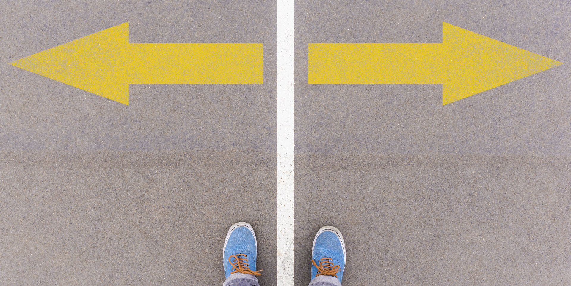 First-person view of a person's feet standing on a gray surface with two large yellow arrows pointing in opposite directions, symbolizing a crossroad or decision-making point.