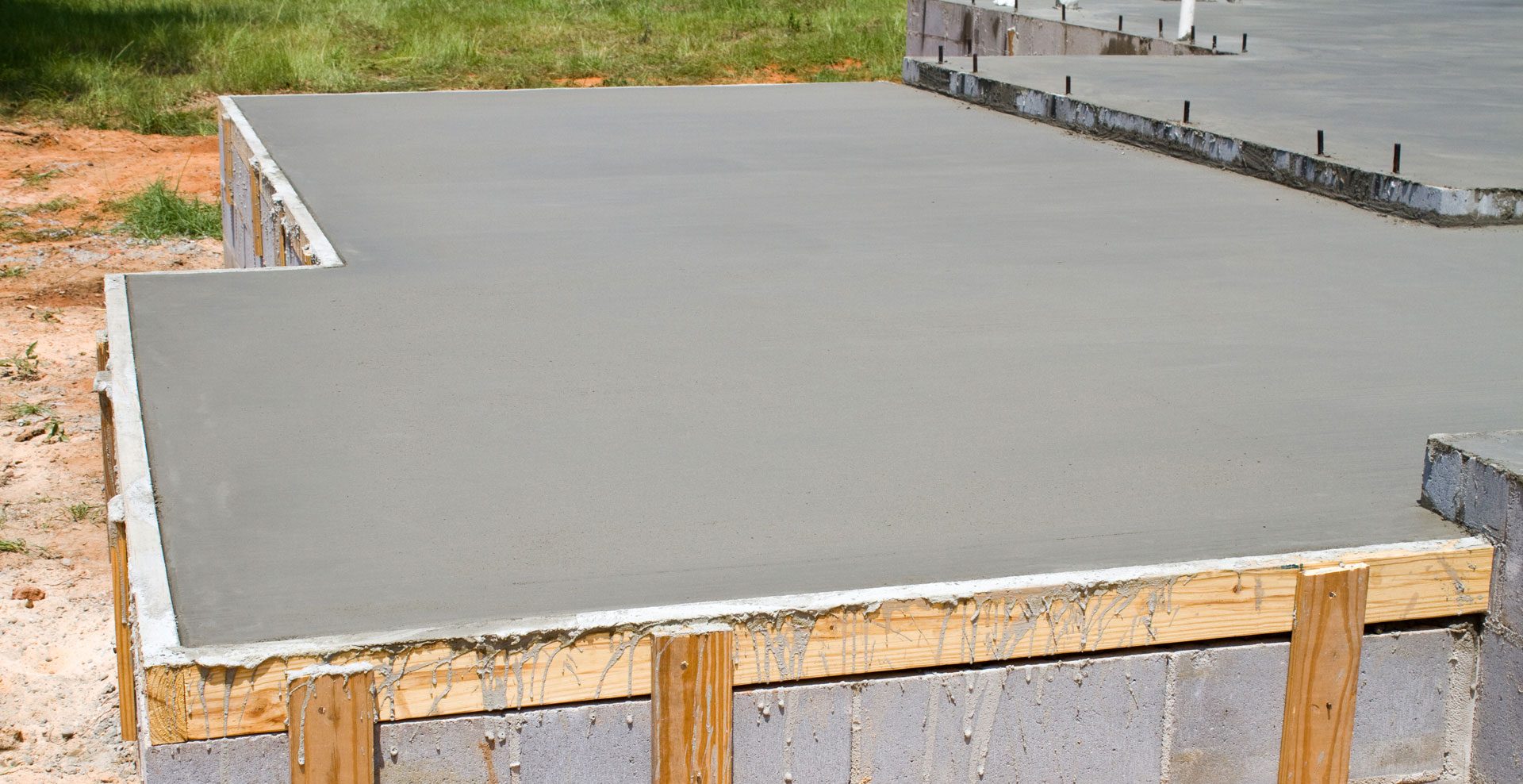A concrete foundation for a building under construction, with exposed steel rebar and wooden formwork still in place around the edges, set against a backdrop of a grassy area.