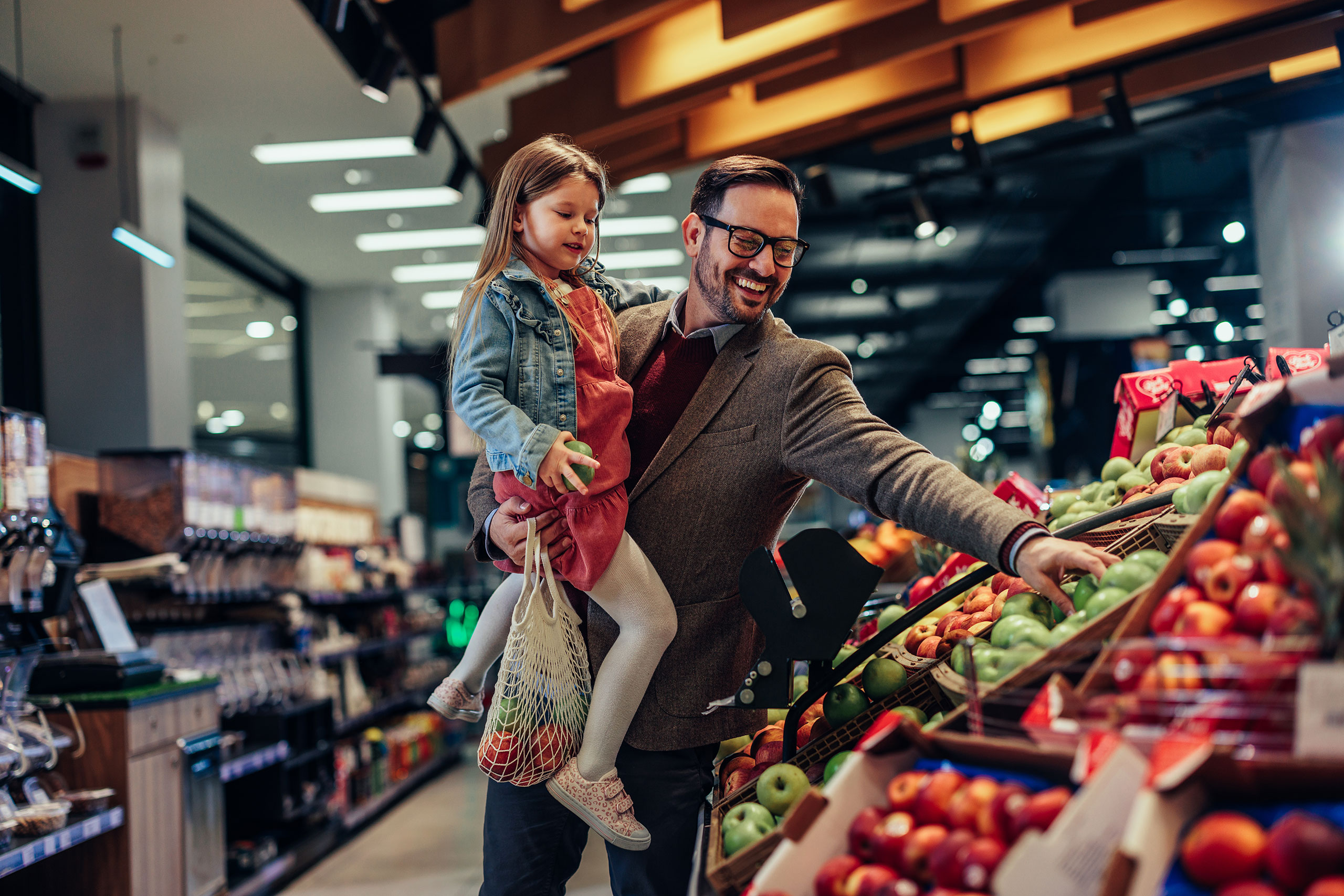 This image shows a father and his young daughter shopping for fresh produce in a grocery store. The father, wearing a blazer and glasses, is holding his daughter while reaching for apples. The daughter, dressed in a denim jacket and pink dress, is holding a green apple and carrying a reusable net bag filled with more produce. They both appear happy and engaged in their shopping experience. The store is well-lit, with neatly arranged fruits and a modern interior design. The image conveys a warm and positive moment of family grocery shopping.
